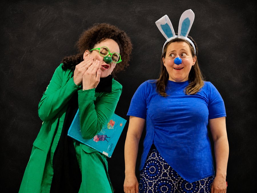 Dos narradoras de cuenta clown en Asturias: Verde (izq, con gafas verdes, nariz verde y un libro bajo el brazo) y Azul (der, con diadema de orejas de conejo y nariz azul) gesticulando en escena.